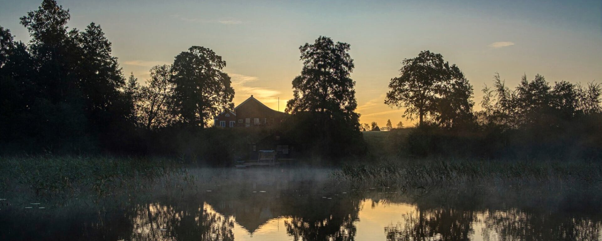 Snokebo Gård vid sjön i Småland i kvällsljus, med dimma över vattnet och omgiven av träd