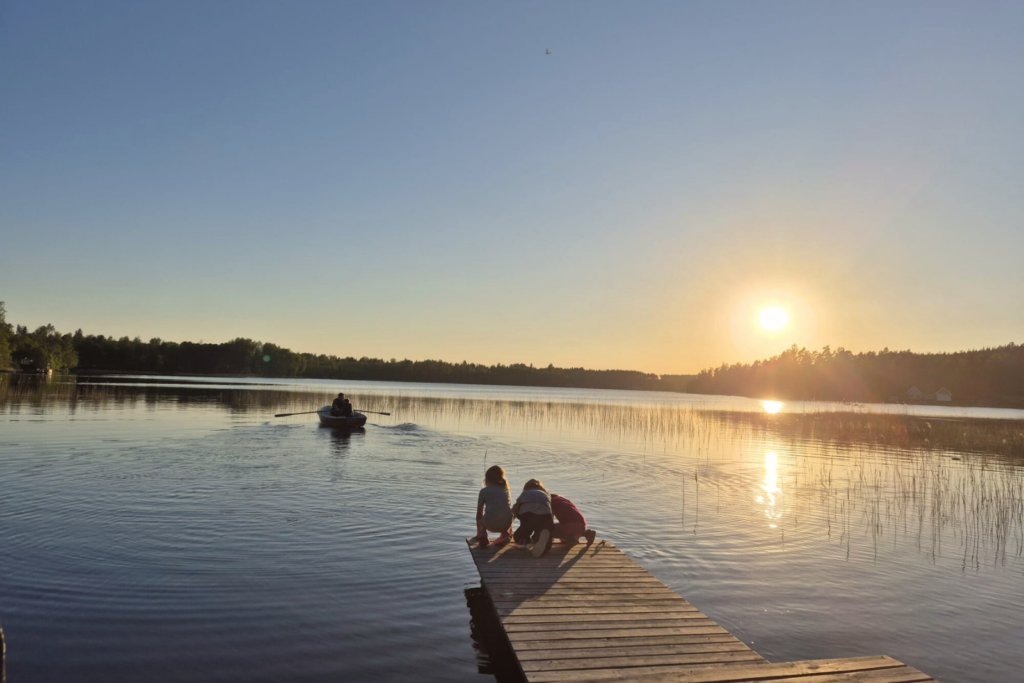 Badplats med brygga vid sjön på Snokebo Gård i Småland, barn vid vattnet och en roddbåt i solnedgången